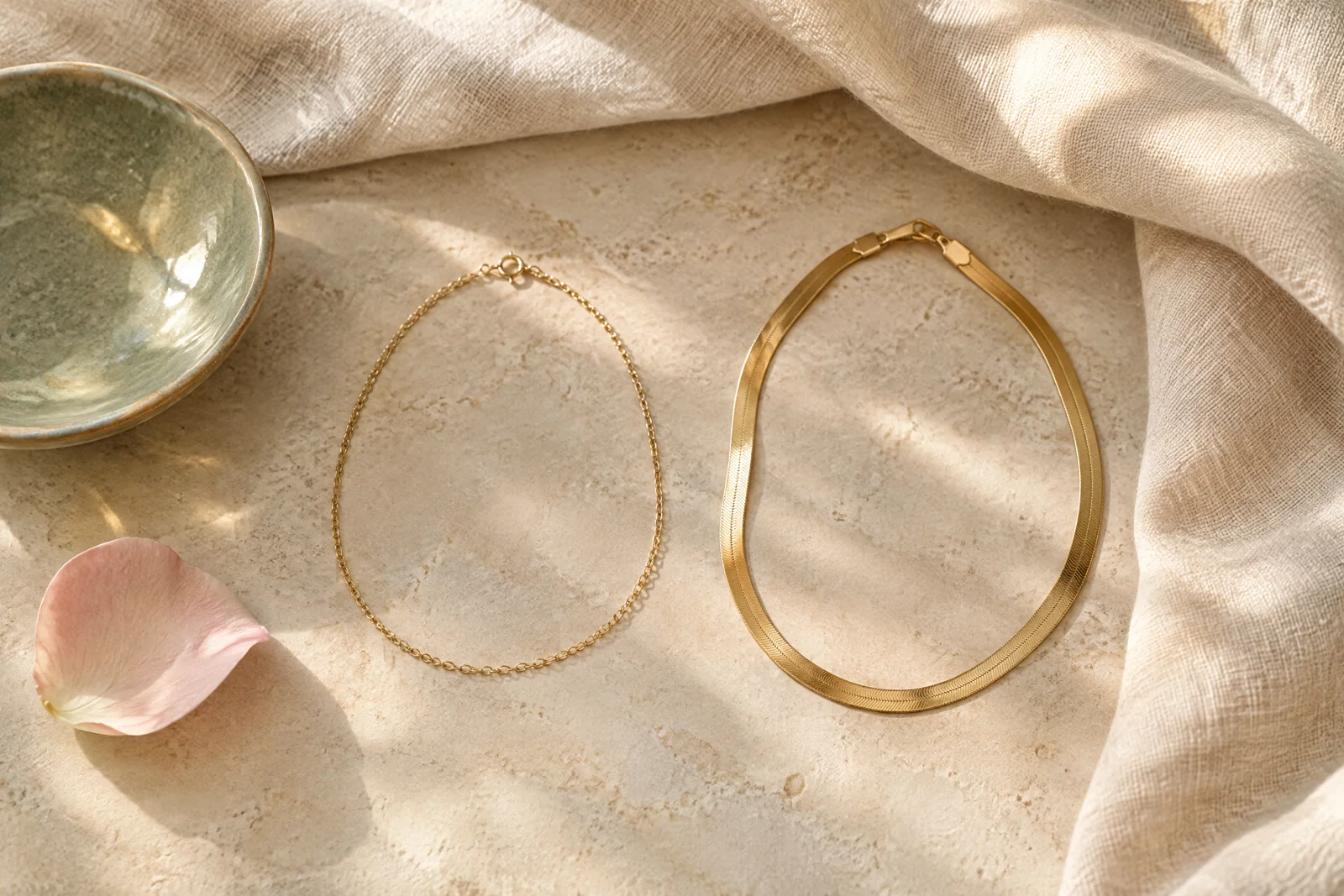 Gold-tone chain and broad band arranged on pale stone with cream linen, a sage ceramic dish, and a blush petal.